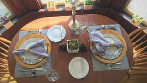 Une table ronde, dressée pour quatre personnes, avec des assiettes à bord doré, des serviettes blanches et des couverts en argent. De petites plantes en pot sont posées sur le rebord de la fenêtre, et un cône de pin sert de centre de table.