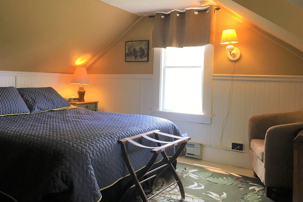 A bedroom with a bed, a chair, a window with curtains, a lamp, and a radiator.