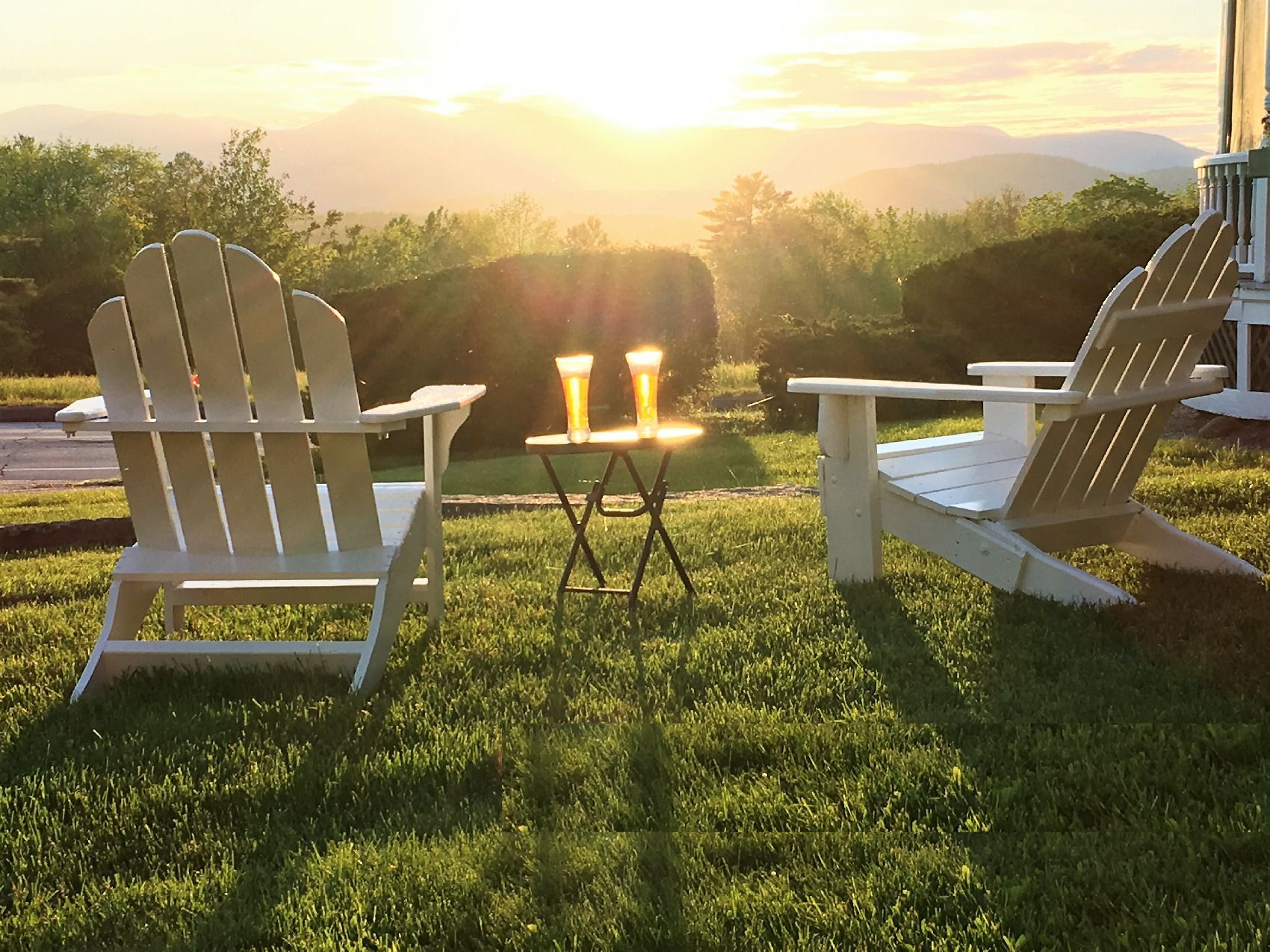 Duas cadeiras Adirondack com uma pequena mesa e duas velas no meio, colocadas em um gramado com vista panorâmica para montanhas e um pôr do sol ao fundo.