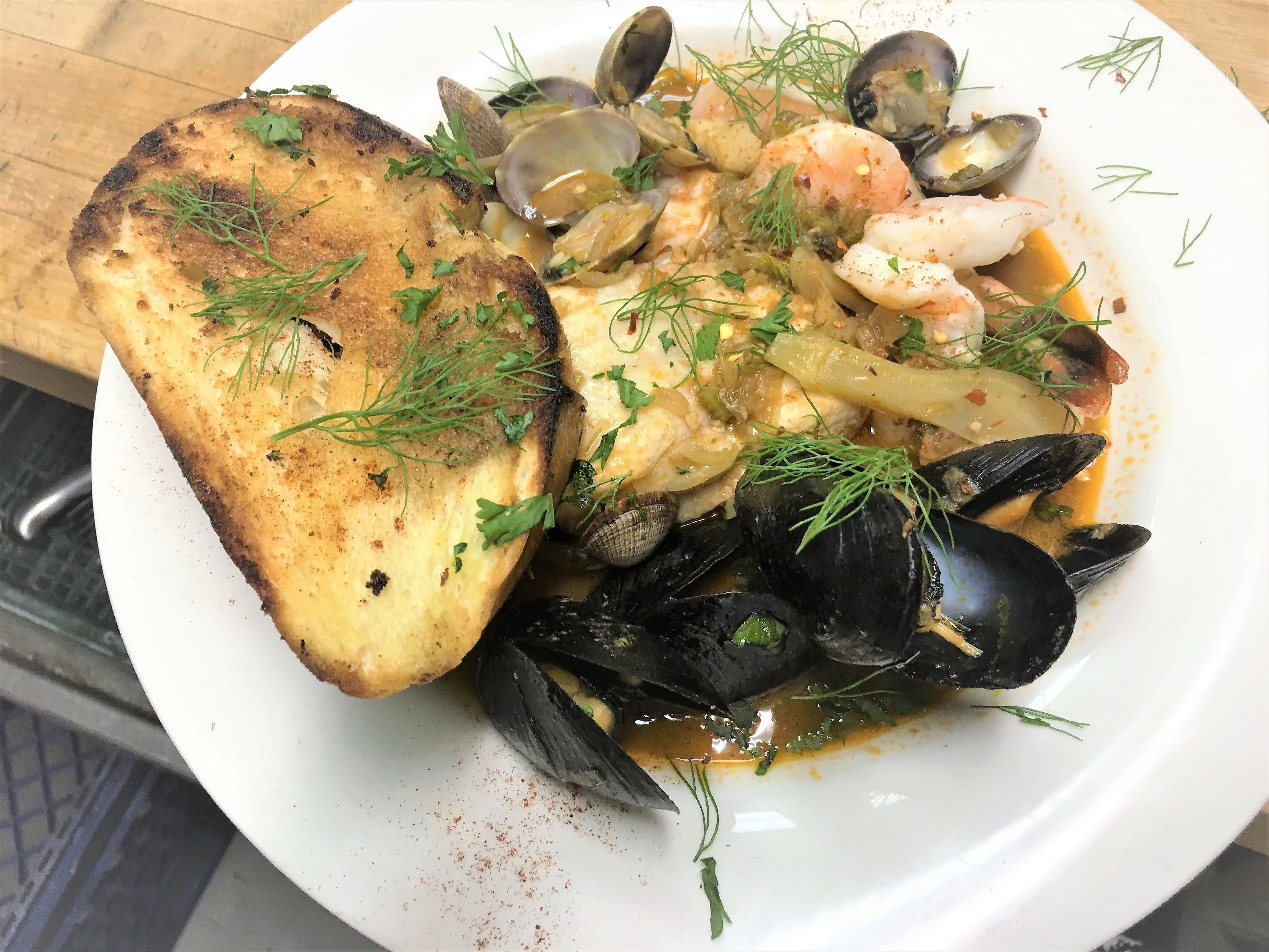 A seafood dish with mussels, clams, shrimp, and a piece of grilled bread, garnished with herbs.
