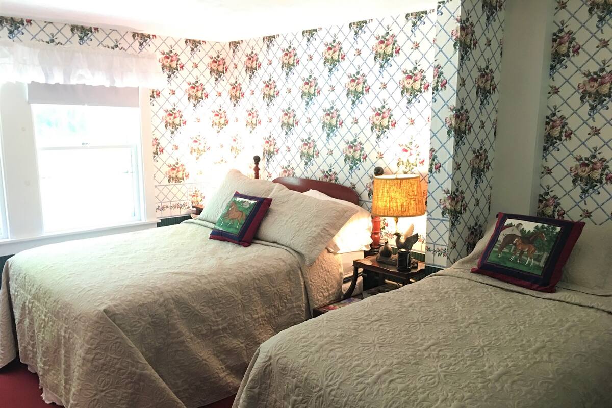 A bedroom with two beds, floral wallpaper, a window with a sheer curtain, and a nightstand with a lamp.