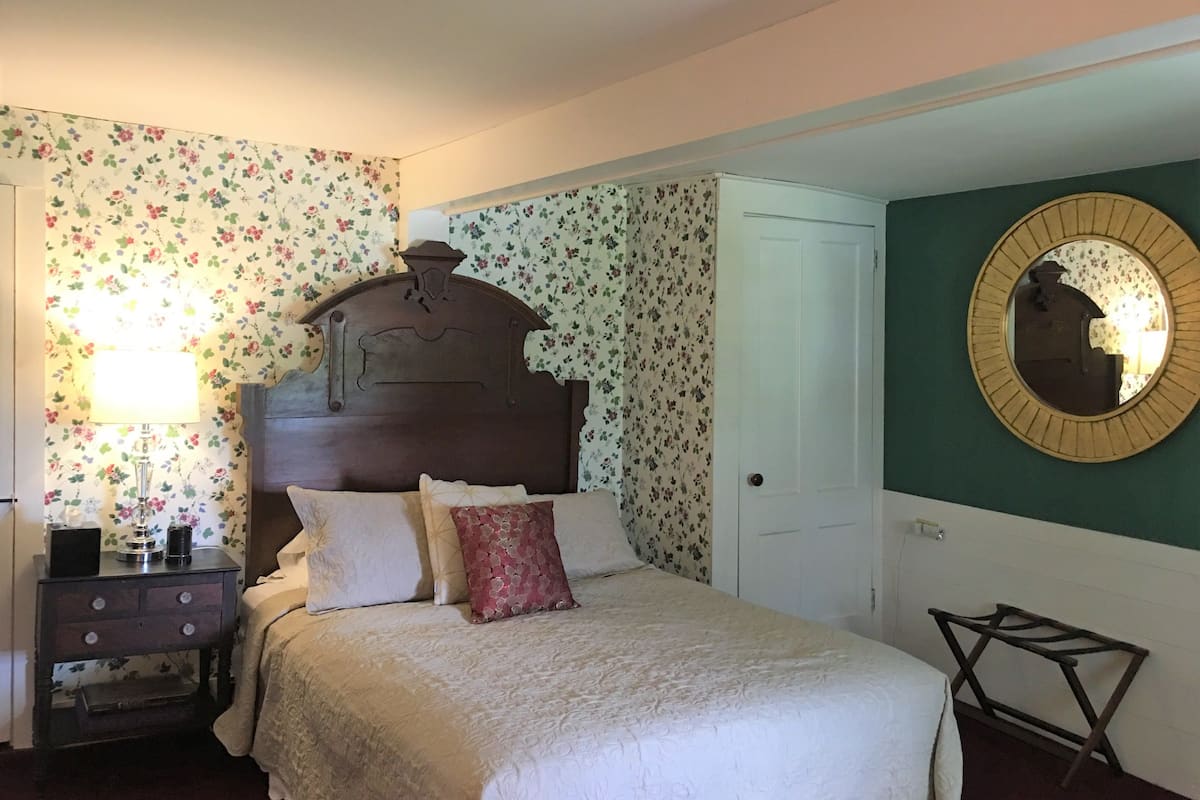 A bedroom with a wooden headboard, floral wallpaper, a bedside table with a lamp, and a round mirror on the wall.