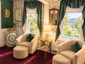 A cozy room with two armchairs, a small table, and a lamp. There are two windows with curtains, a potted plant, and a mirror on the wall. - Center Lovell Inn (Lovell)