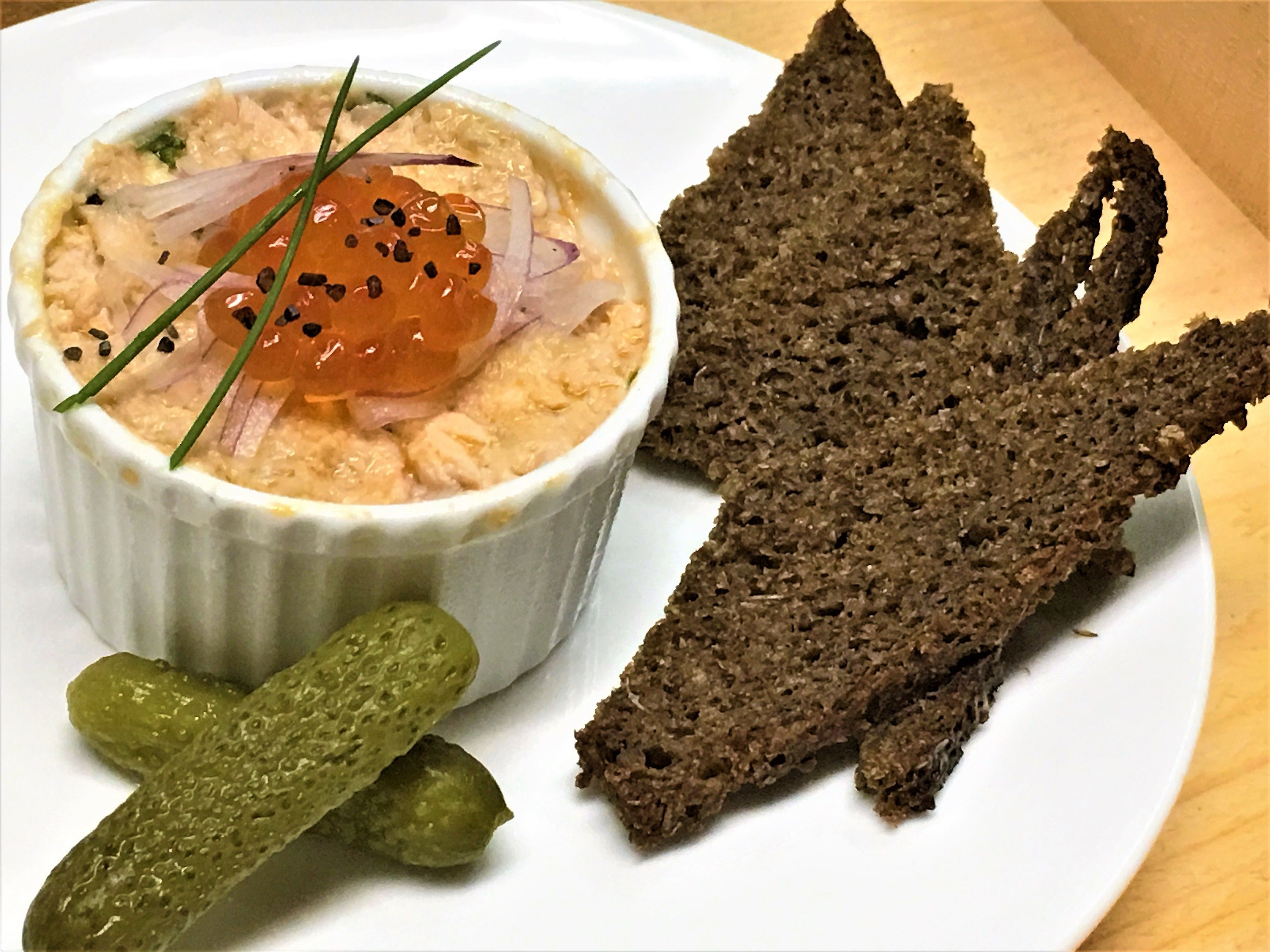 A plate with a bowl of dip garnished with chives and black pepper, accompanied by two pickles and a piece of dark rye bread.