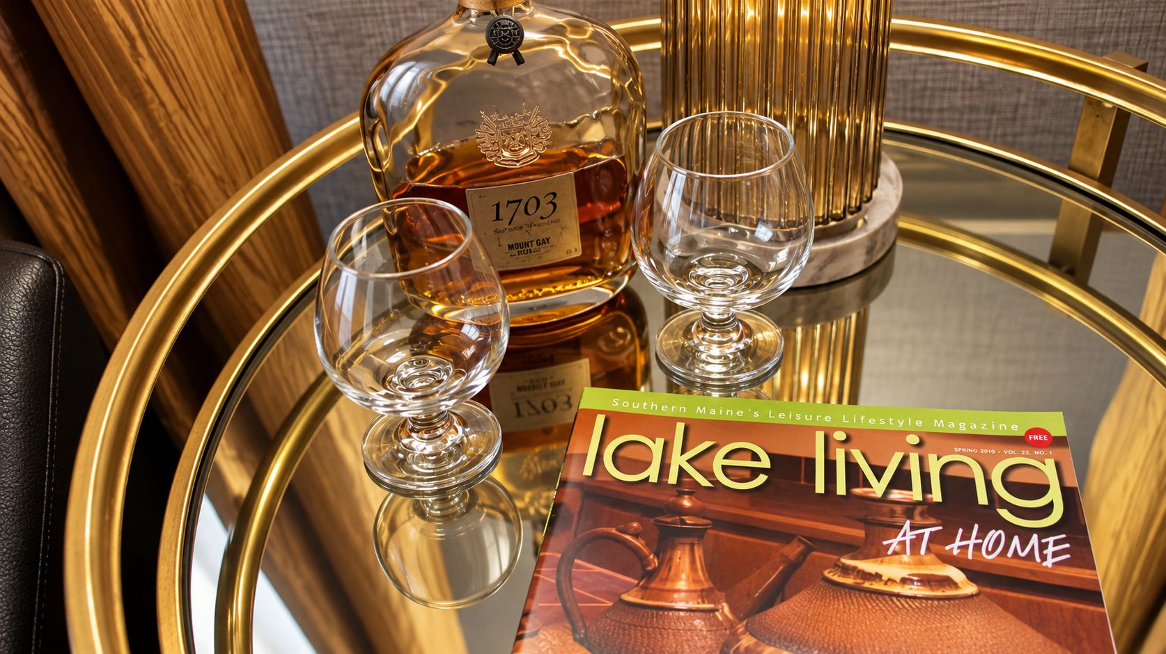 A bottle of Mount Gay whiskey, two glasses, and a magazine titled 'Lake living at home' on a reflective glass table.