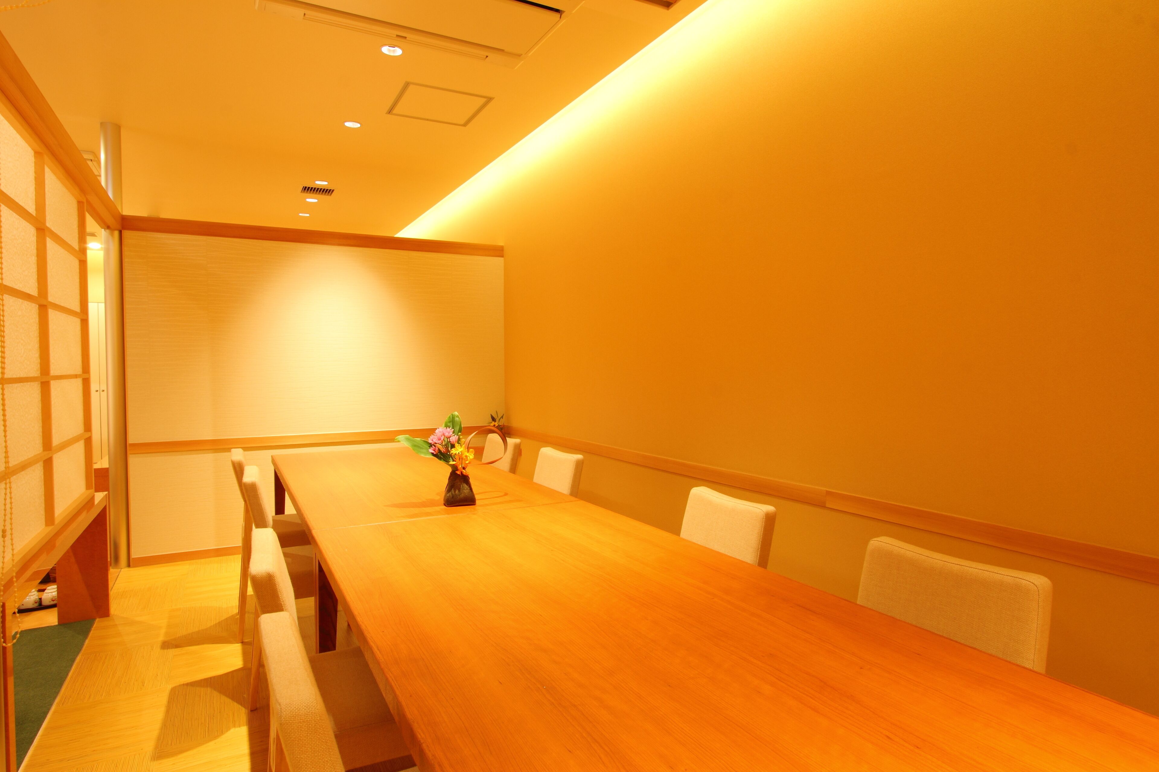 Un salón de conferencias con una larga mesa de madera, sillas blancas y un pequeño jarrón con flores.