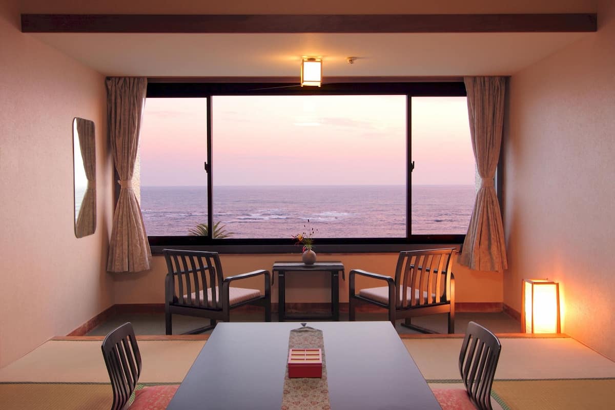 A room with a view of the ocean, a table with chairs, a small table with a vase, and a standing lamp.
