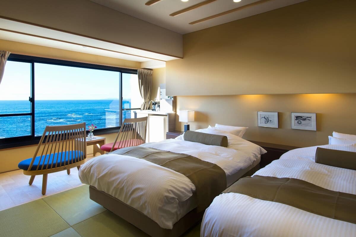 A hotel room with two beds, a desk, a chair, and a large window offering a sea view.