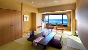 Quarto com piso de tatame, uma mesa baixa com um tapete roxo e branco, duas cadeiras de madeira com almofadas, uma janela grande com vista para o oceano e um guarda-roupa.