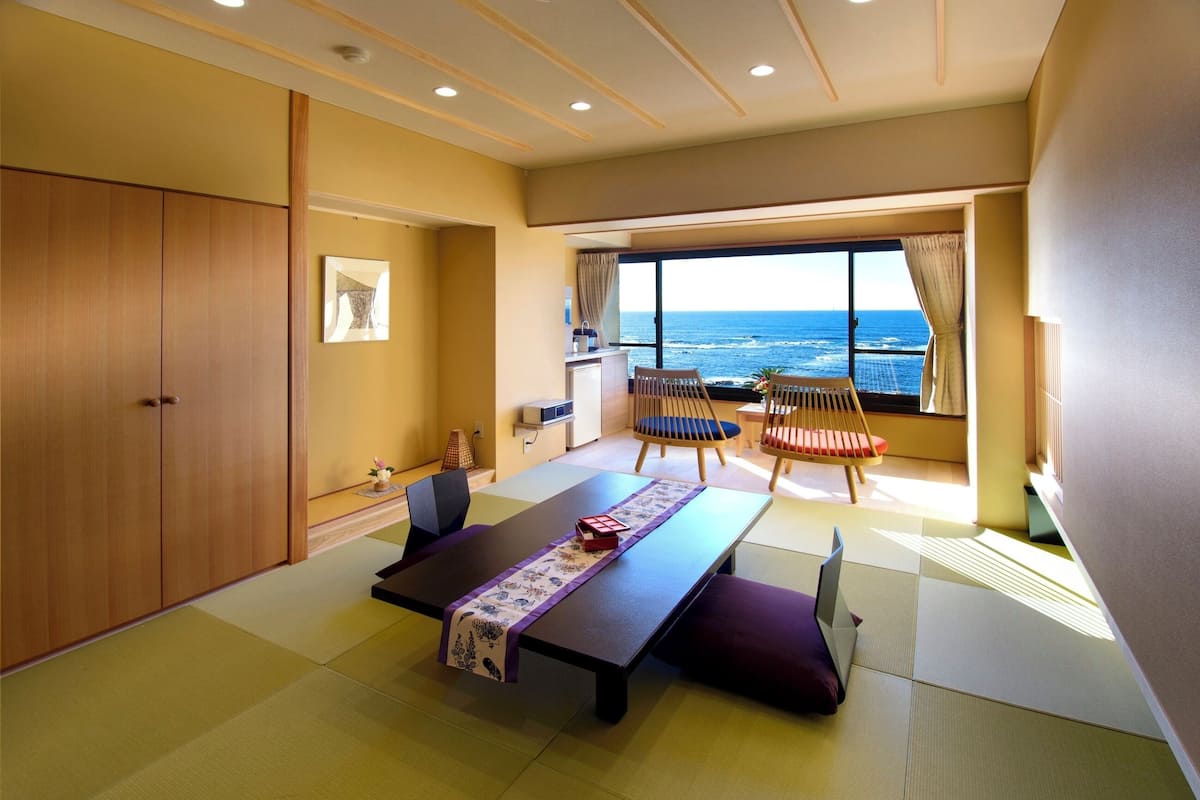 A room with tatami flooring, a low table with a purple and white mat, two wooden chairs with cushions, a large window with a view of the ocean, and a wardrobe.