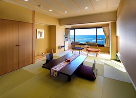 Una habitación con suelo de tatami, una mesa baja con esterilla morada y blanca, dos sillas de madera con cojines, un ventanal con vista al océano y un armario.
