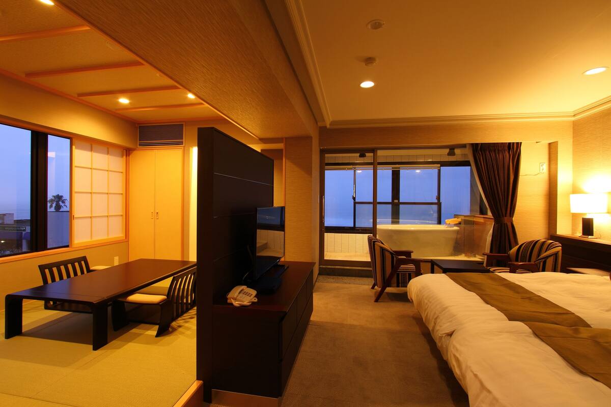 A hotel room with a large bed, a desk with a chair, a TV, and a window with a view of the city.