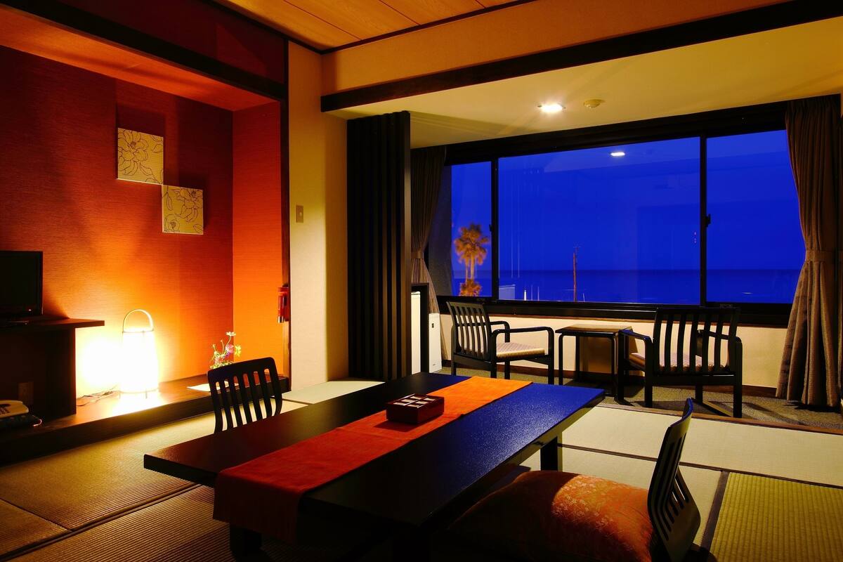 A hotel room with a dining table, chairs, a TV, and a large window with a view of the sea.