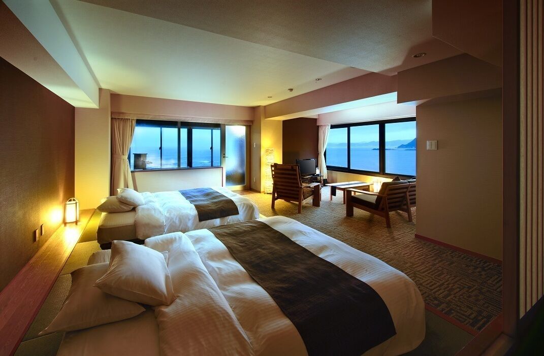 A hotel room with two beds, a desk, a chair, and large windows offering a view of the sea.