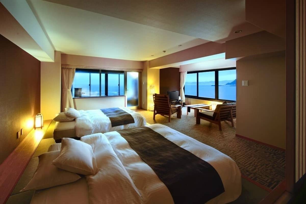 A hotel room with two beds, a desk, a chair, and large windows offering a view of the sea.