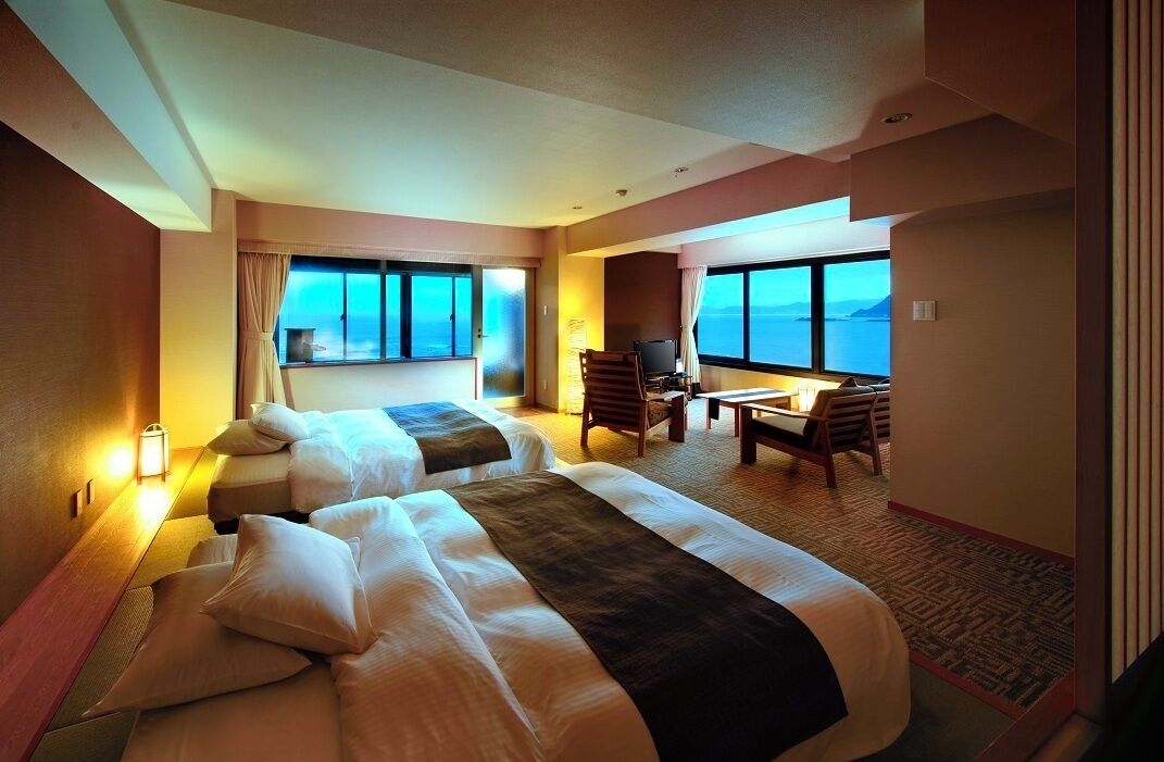 A hotel room with two beds, a desk, a chair, and large windows with a view.