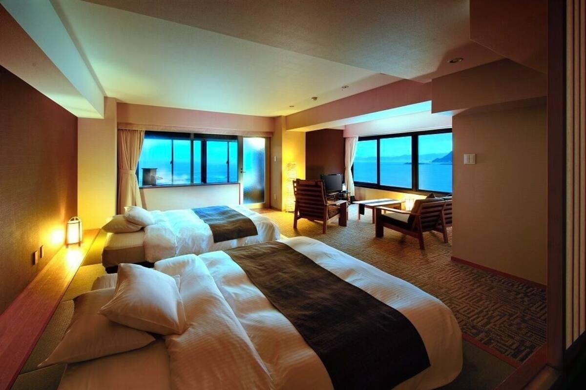 A hotel room with two beds, a desk, a chair, and large windows with a view.