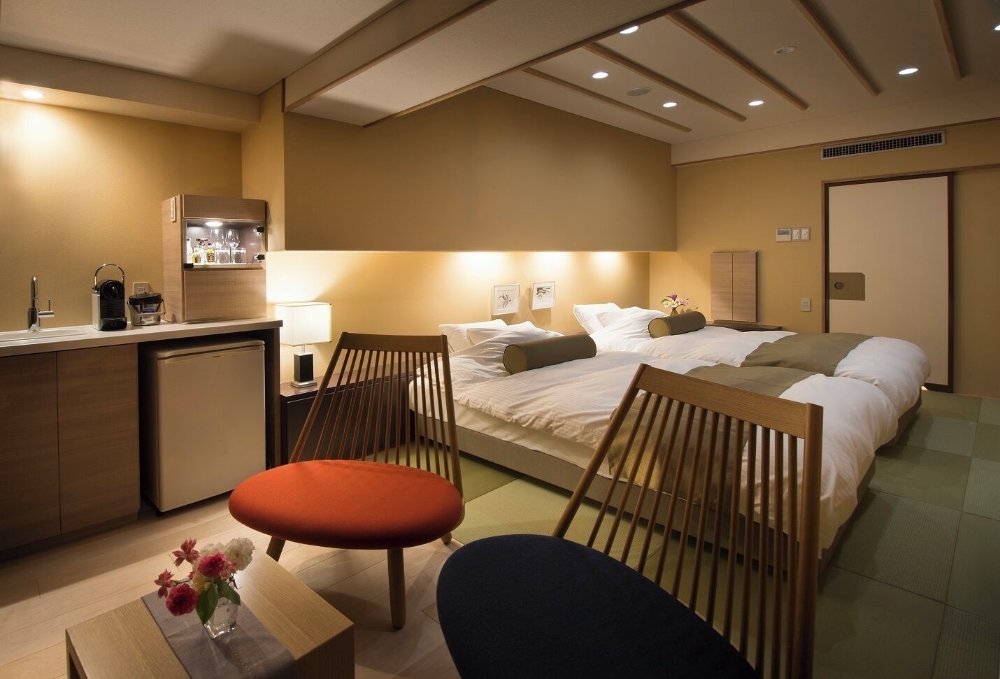 A hotel room with a bed, two chairs, a small table with a vase of flowers, a kitchenette, and a bathroom.