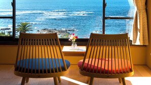 Duas cadeiras de madeira com almofadas azuis e vermelhas voltadas para uma janela com vista para o oceano.