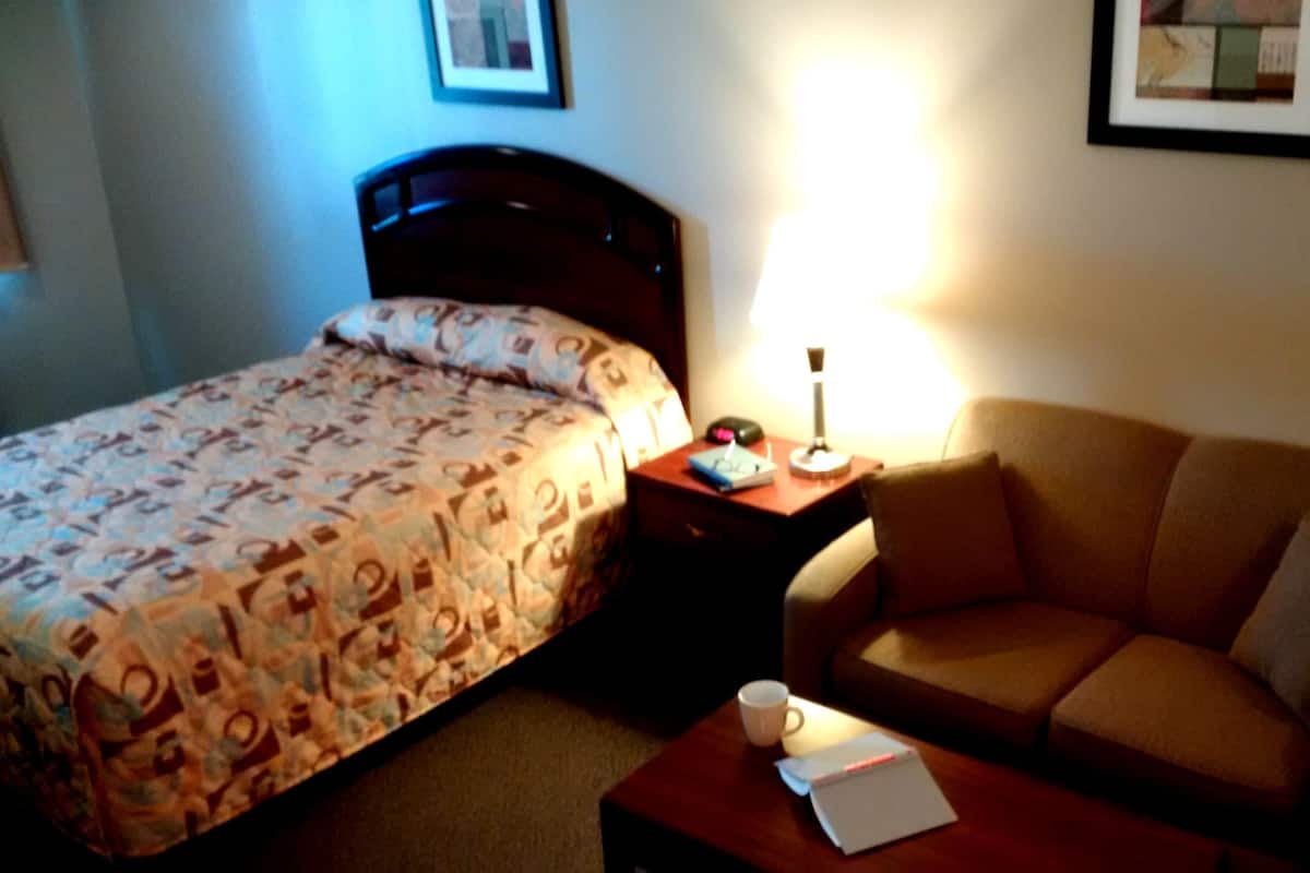 A hotel room with a bed, a couch, a nightstand with a lamp, and a coffee table with a cup and a notebook.