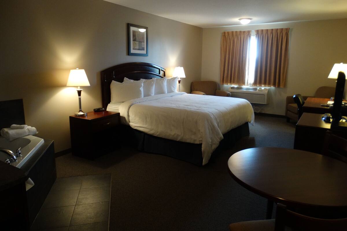 A hotel room with a large bed, two bedside tables with lamps, a desk with a chair, a small table, and a window with curtains.