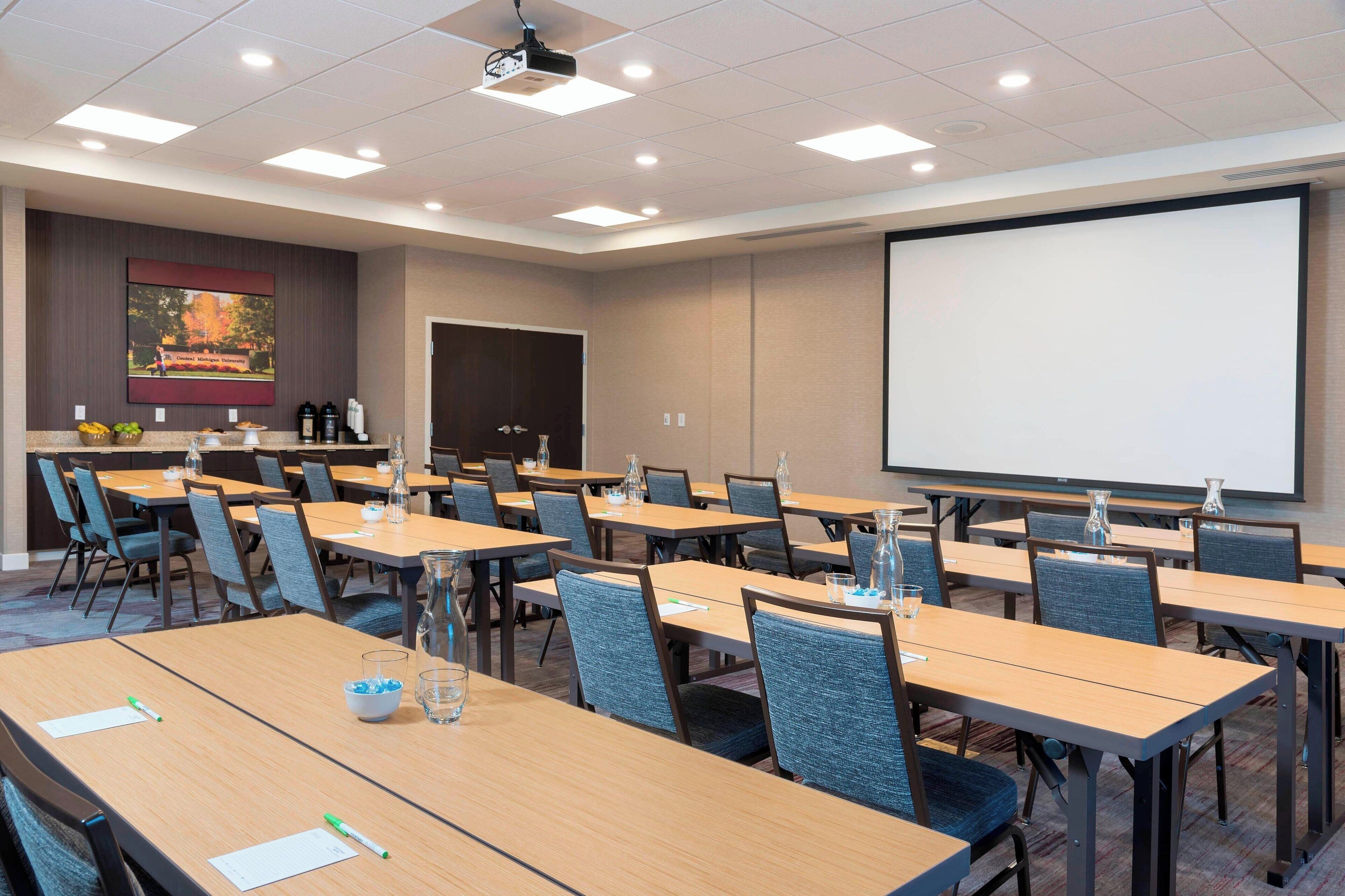 A conference room with multiple tables, chairs, a projector screen, and a presentation screen.