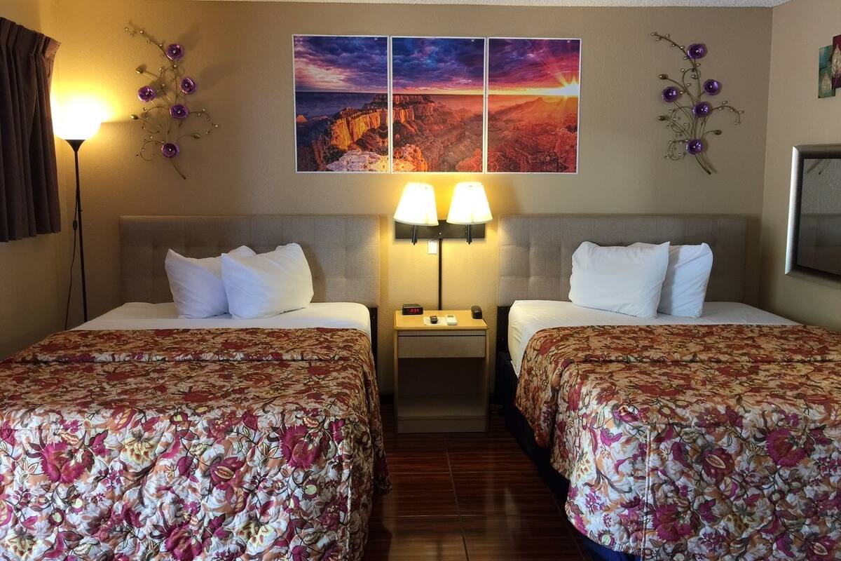 A hotel room with two beds, floral bedspreads, a nightstand with lamps, and a wall with three framed pictures.
