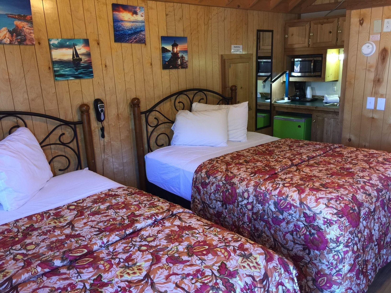 A room with two beds, wooden walls, and a kitchen area visible through an open door.