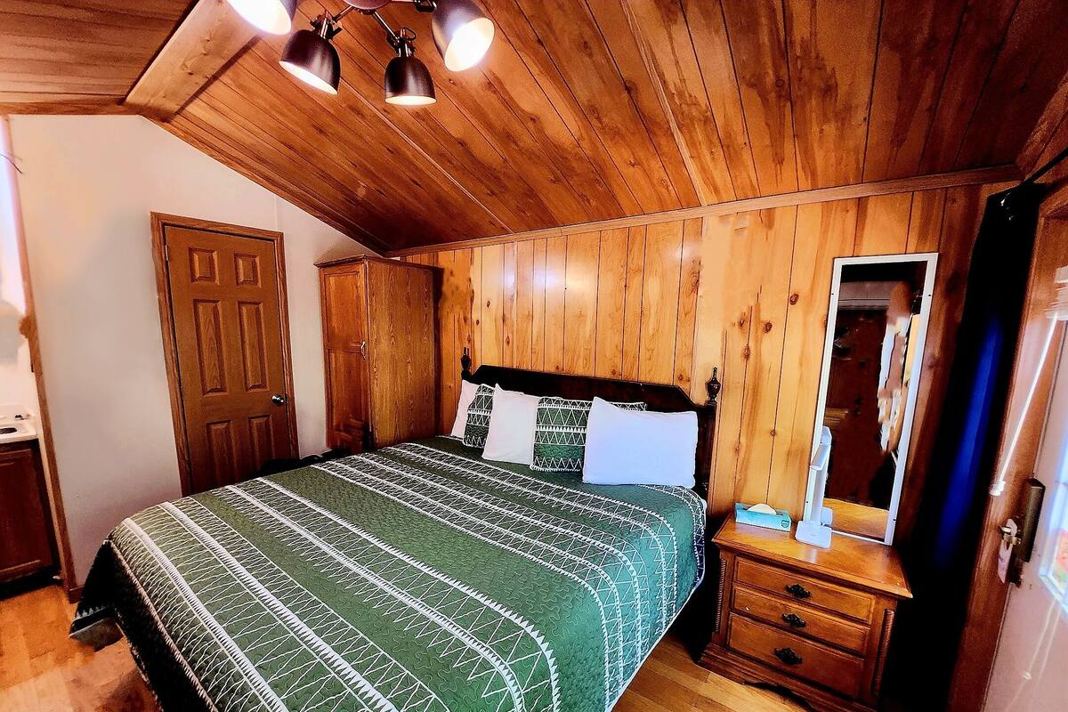 A wooden-paneled bedroom with a bed, a wooden nightstand, a mirror, and a door.
