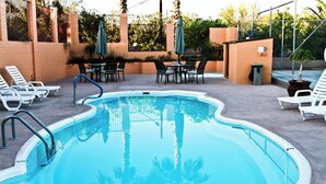 Une piscine entourée d’une clôture et de verdure, avec des chaises longues et des tables à proximité.