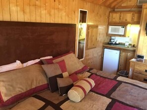A wooden cabin interior with a bed, pillows, a small kitchenette, and a wooden ceiling.