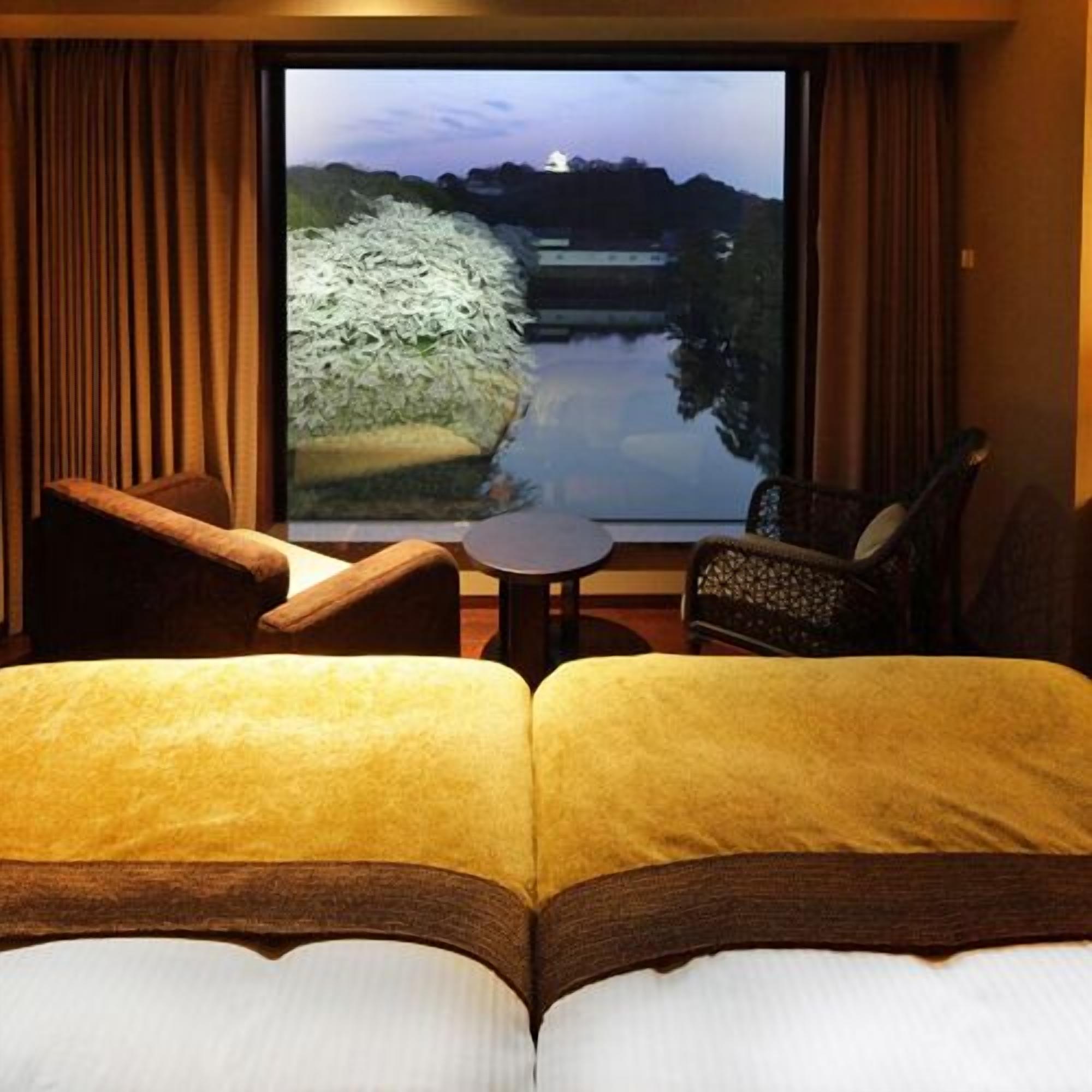 Habitación de hotel con cama, dos sillas, una mesita pequeña y un amplio ventanal con vistas al agua y a las montañas.