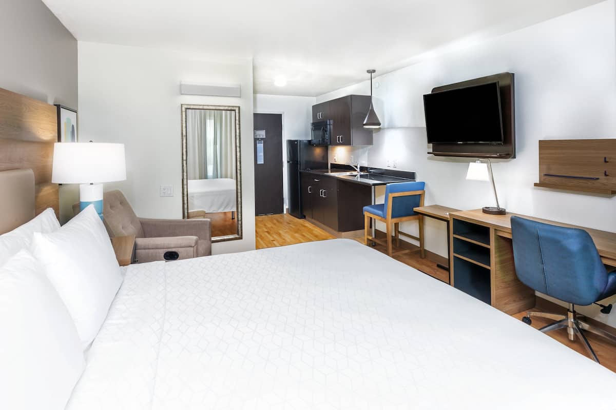 A hotel room with a large bed, a desk with a chair, a TV, and a kitchenette.