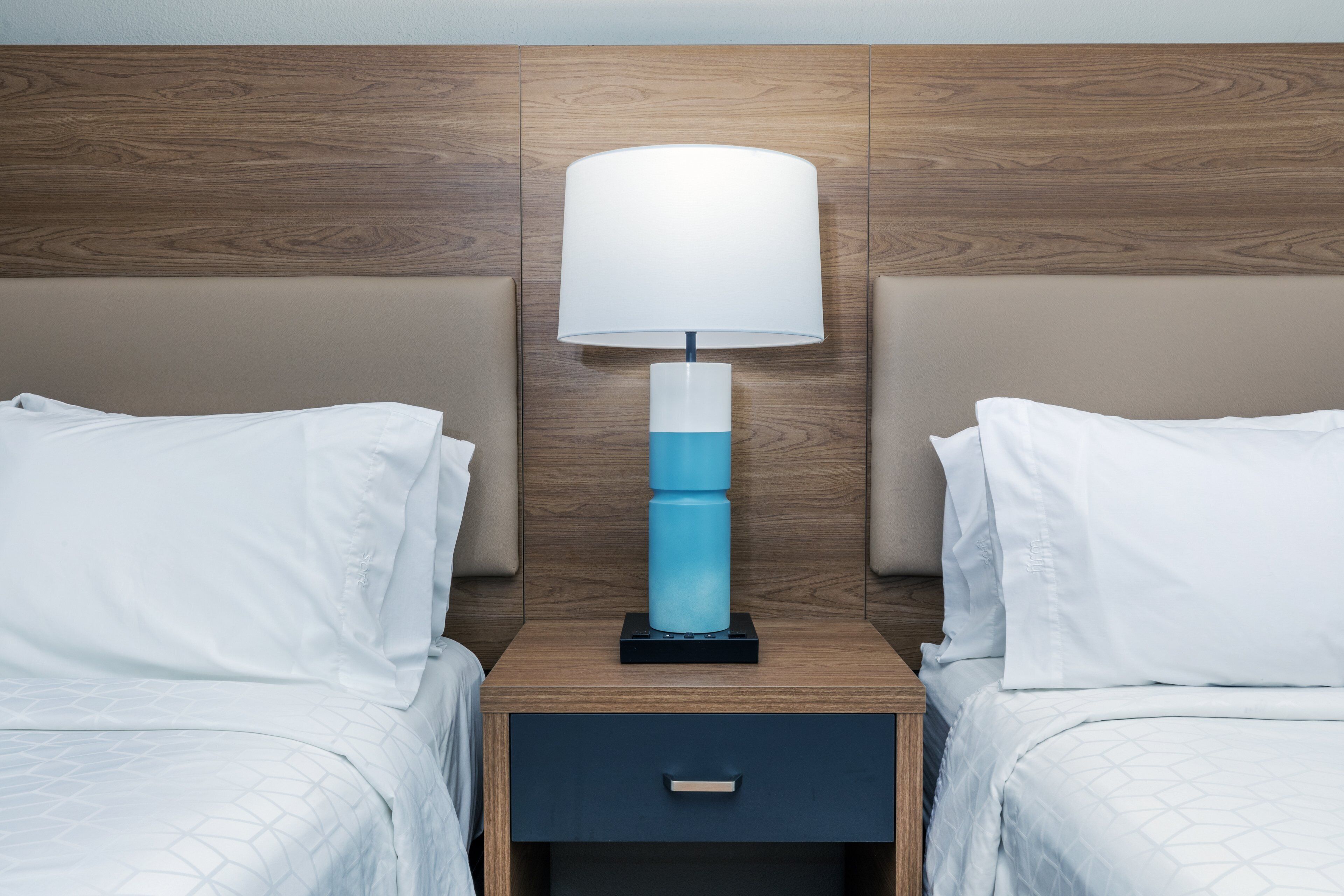 A hotel room with a wooden headboard, a bedside lamp, two beds with white bedding, and a small drawer under the nightstand.
