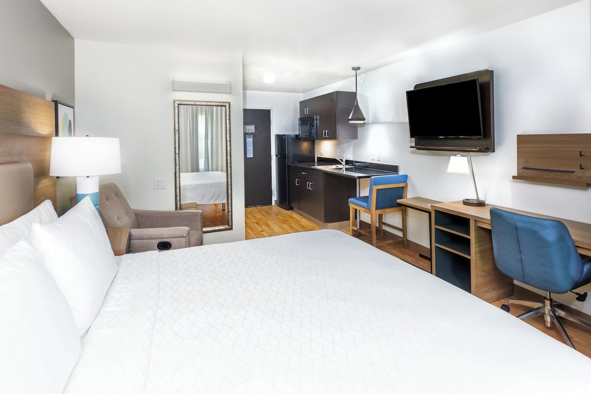 A hotel room with a large bed, a desk with a chair, a TV, and a kitchenette.