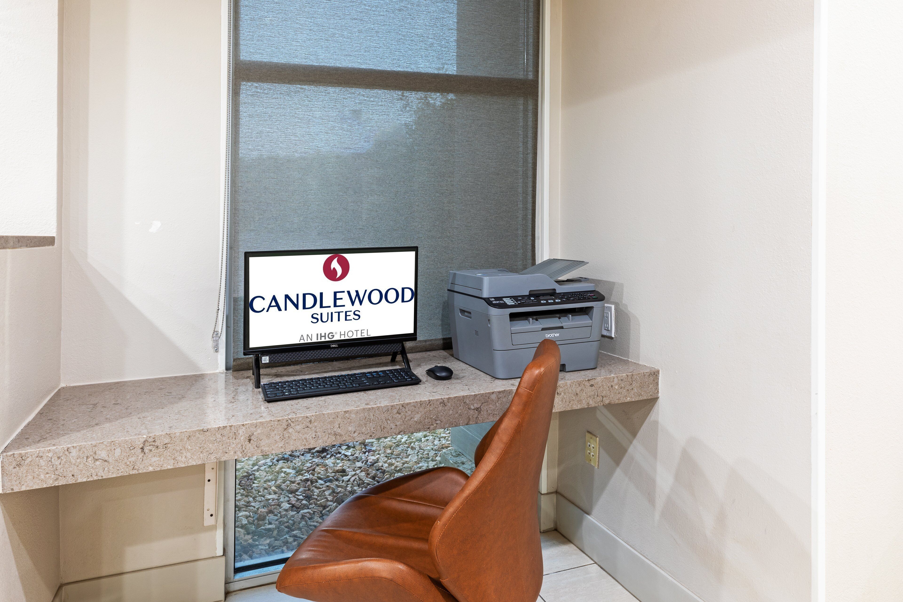 Un escritorio de computadora con un monitor que muestra "CANDLEWOOD SUITES AN IHG HOTEL", una impresora y una silla de cuero marrón.