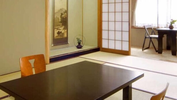 Una habitación japonesa tradicional con suelo de tatami, una mesa baja, una silla, una puerta corredera y una ventana con cortinas.