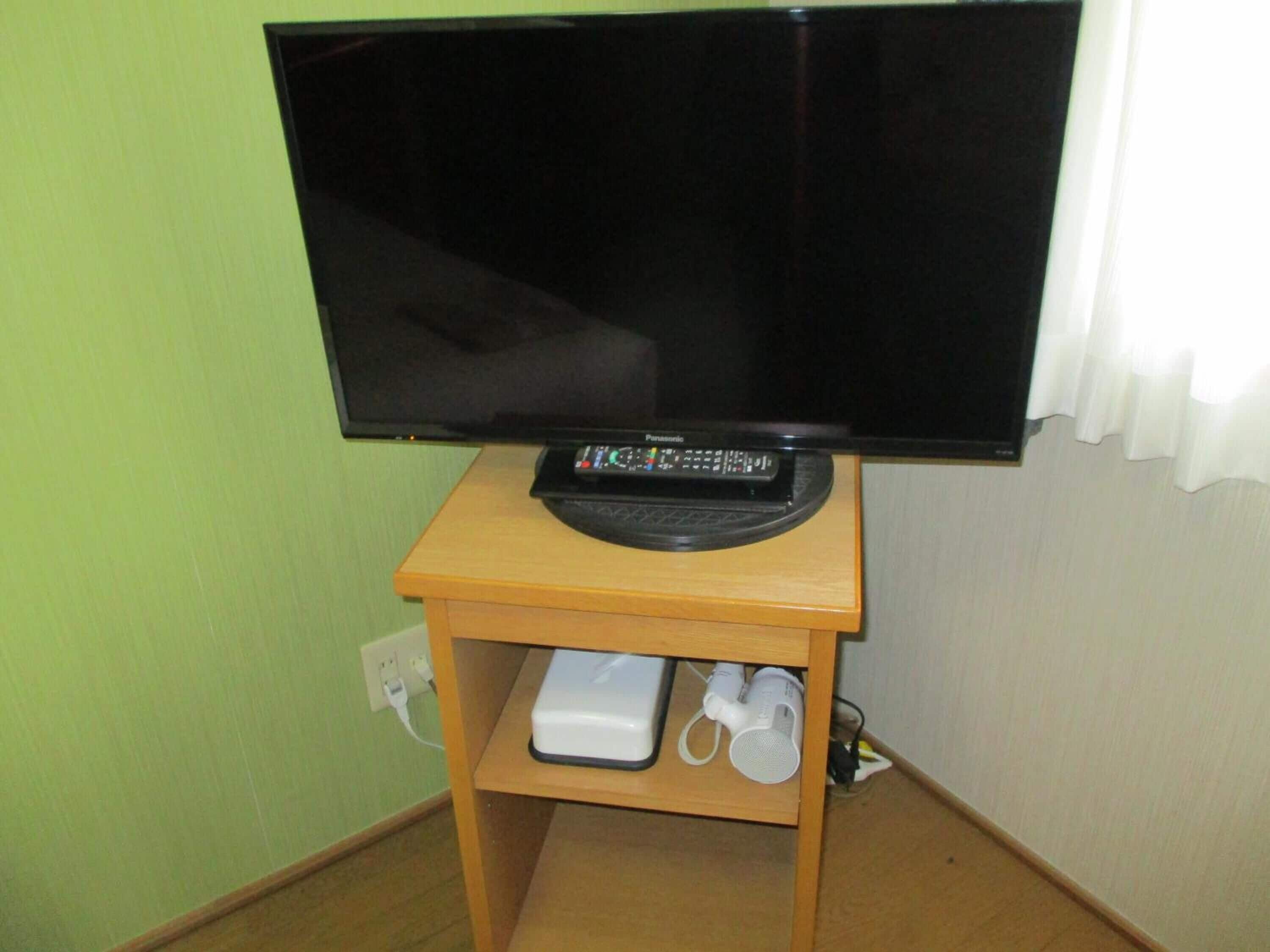 Un televisor sobre un soporte de madera, un mando a distancia, un dispositivo blanco y un altavoz blanco en una estantería.