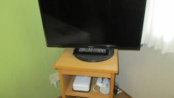 Un televisor sobre un soporte de madera, un mando a distancia, un dispositivo blanco y un altavoz blanco en una estantería.