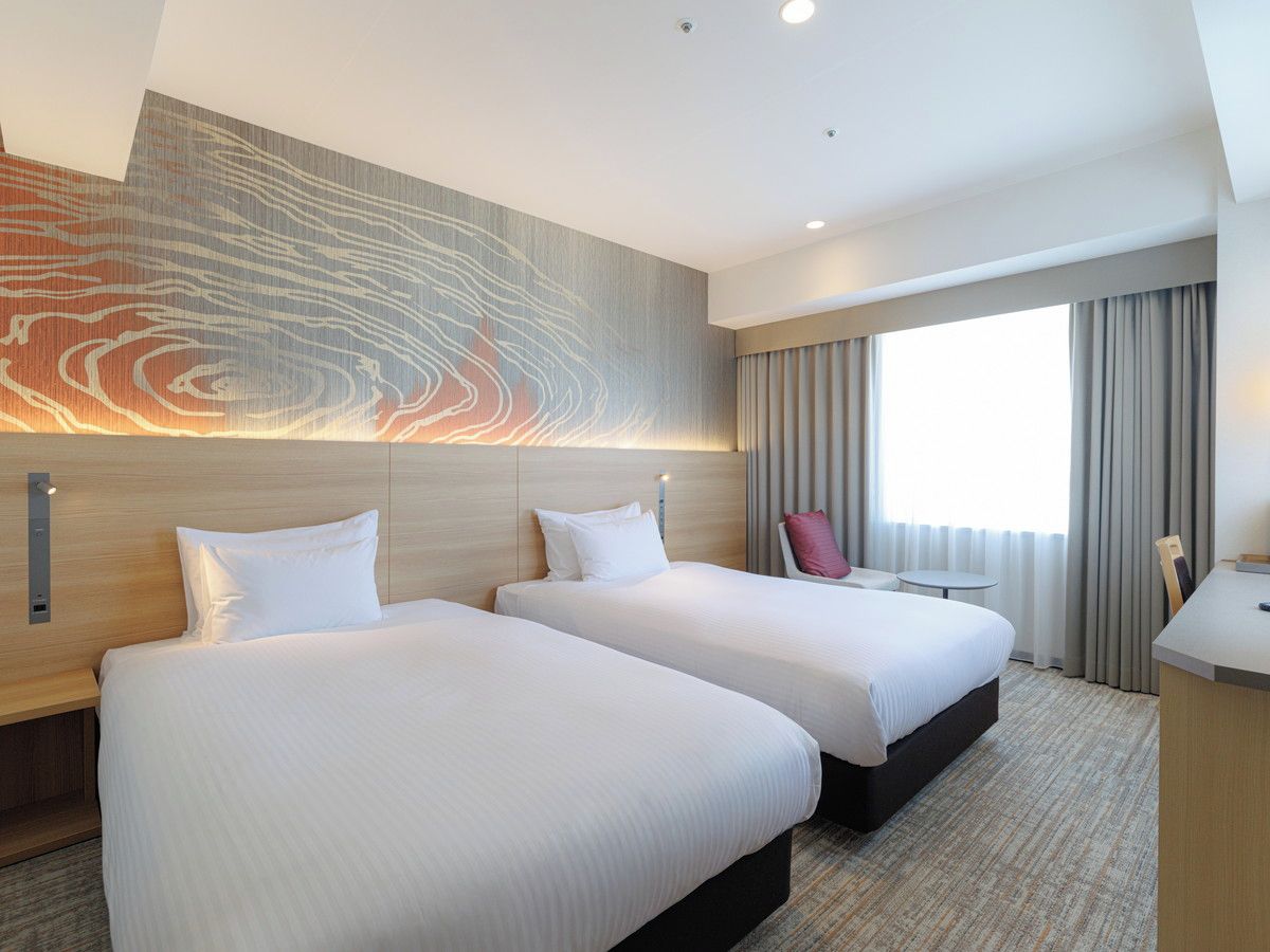 Une chambre d’hôtel avec deux lits, un bureau, une chaise, une grande œuvre d’art abstraite accrochée au mur et une fenêtre avec des rideaux.