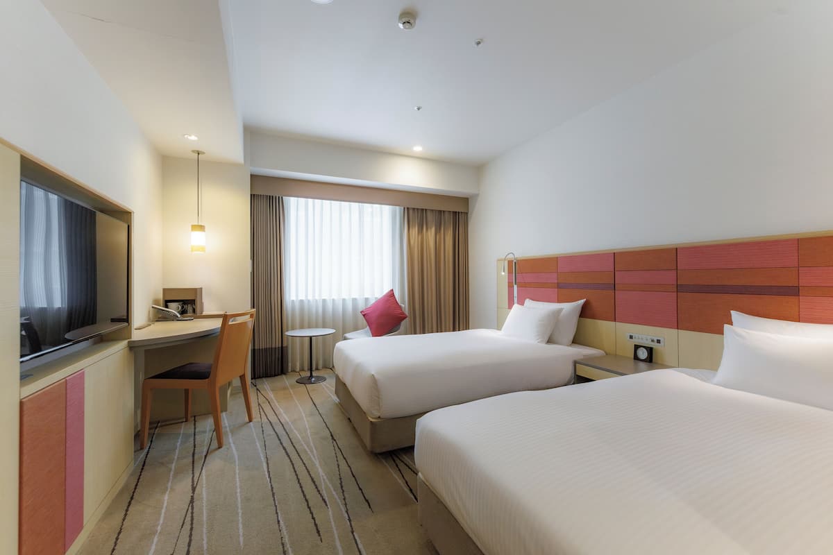 A hotel room with two beds, a desk, a chair, a TV, and a small table with a lamp.