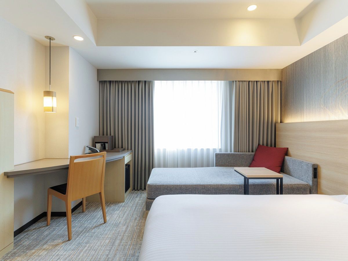 A hotel room with a bed, a desk, a chair, a sofa, and a window with curtains.