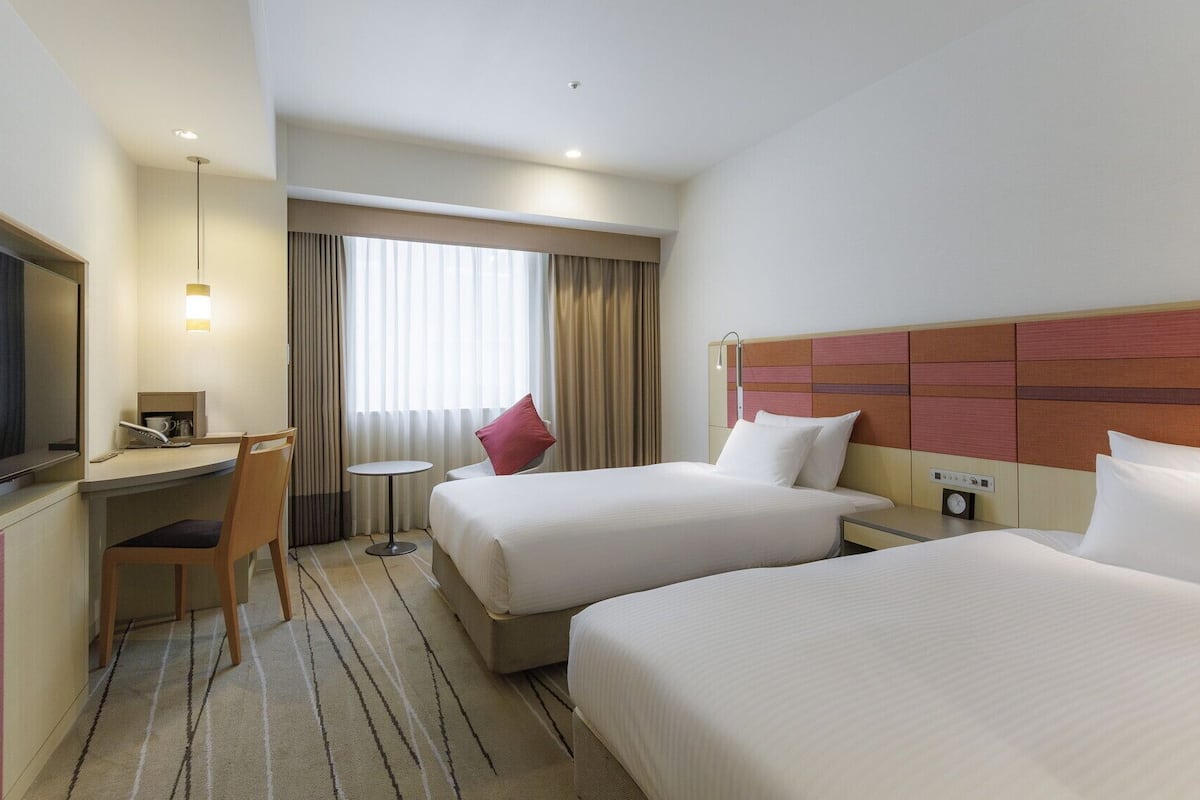 A hotel room with two beds, a desk, a chair, a TV, and a window with curtains.