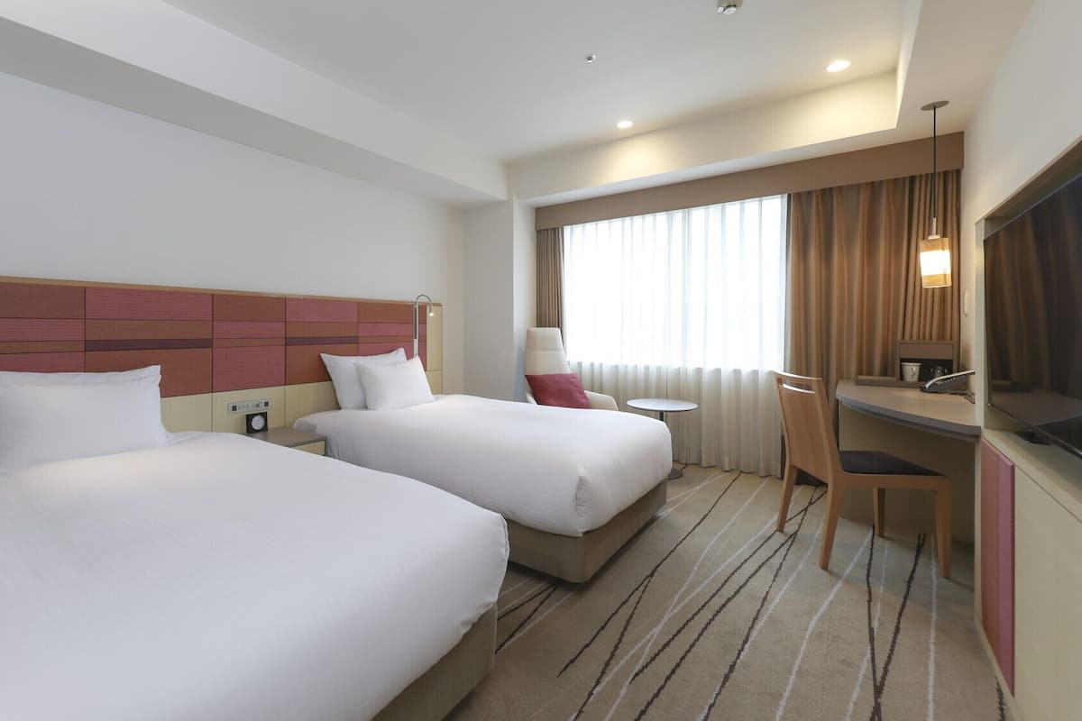 A hotel room with two beds, a desk, a chair, a TV, and a window with curtains.