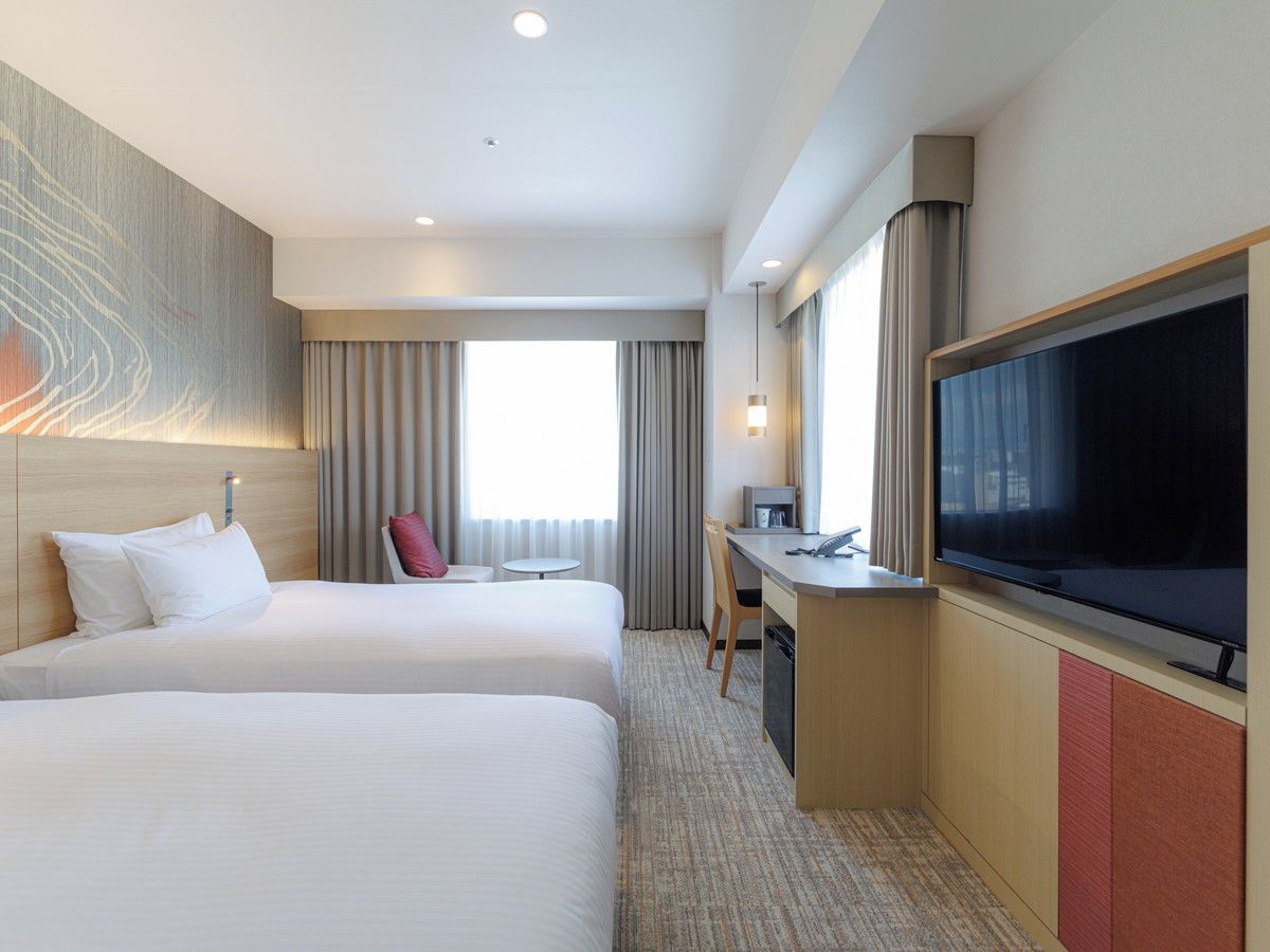 A hotel room with two beds, a desk, a television, and a large window.
