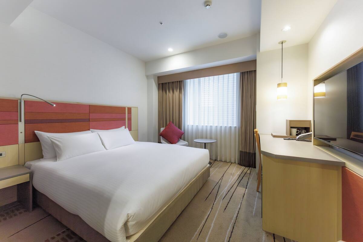 A hotel room with a large bed, a desk, a chair, a small table, a mirror, and a window with curtains.