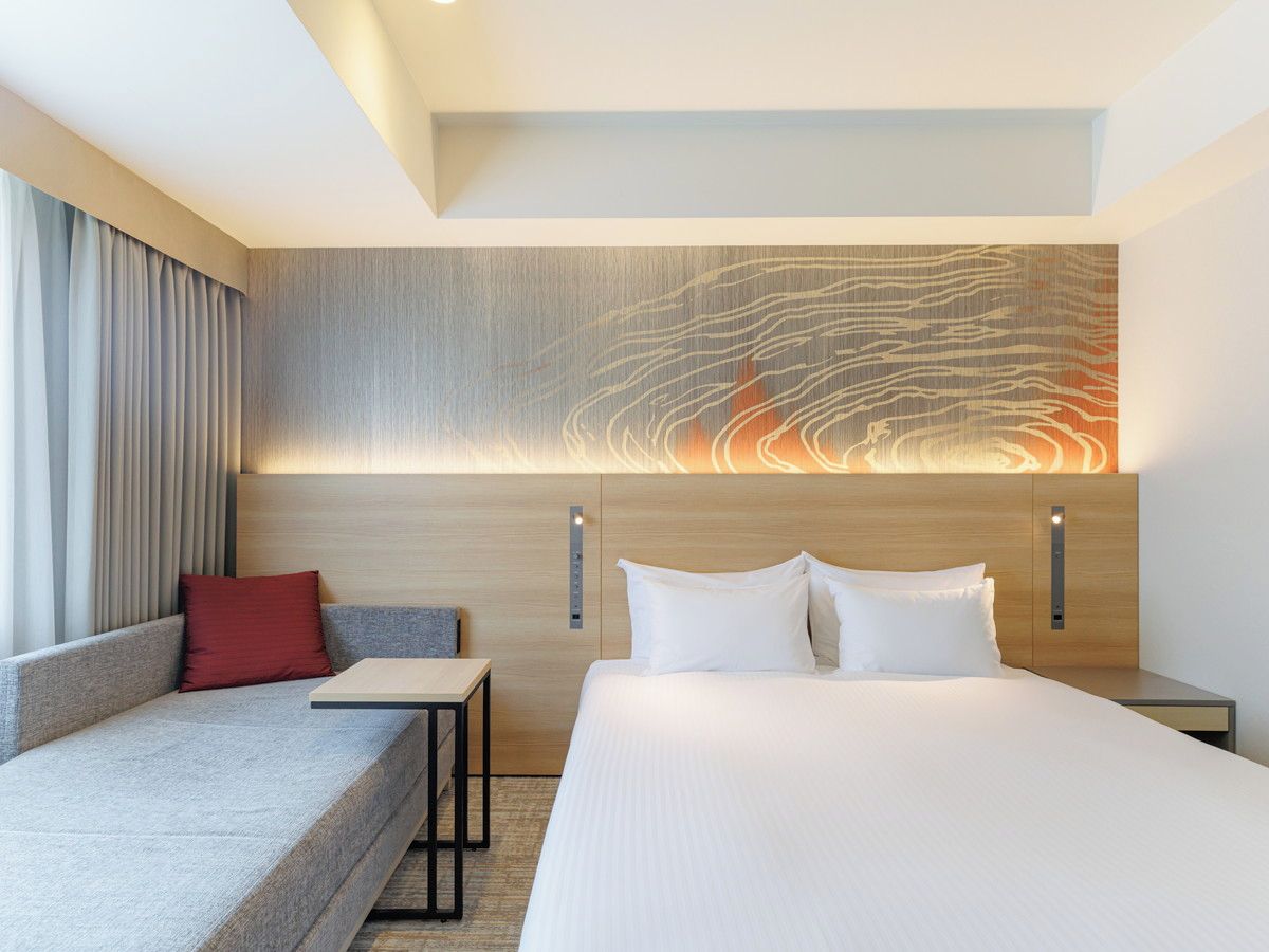 A modern hotel room with a bed, a sofa, a small table, and a wall-mounted artwork.