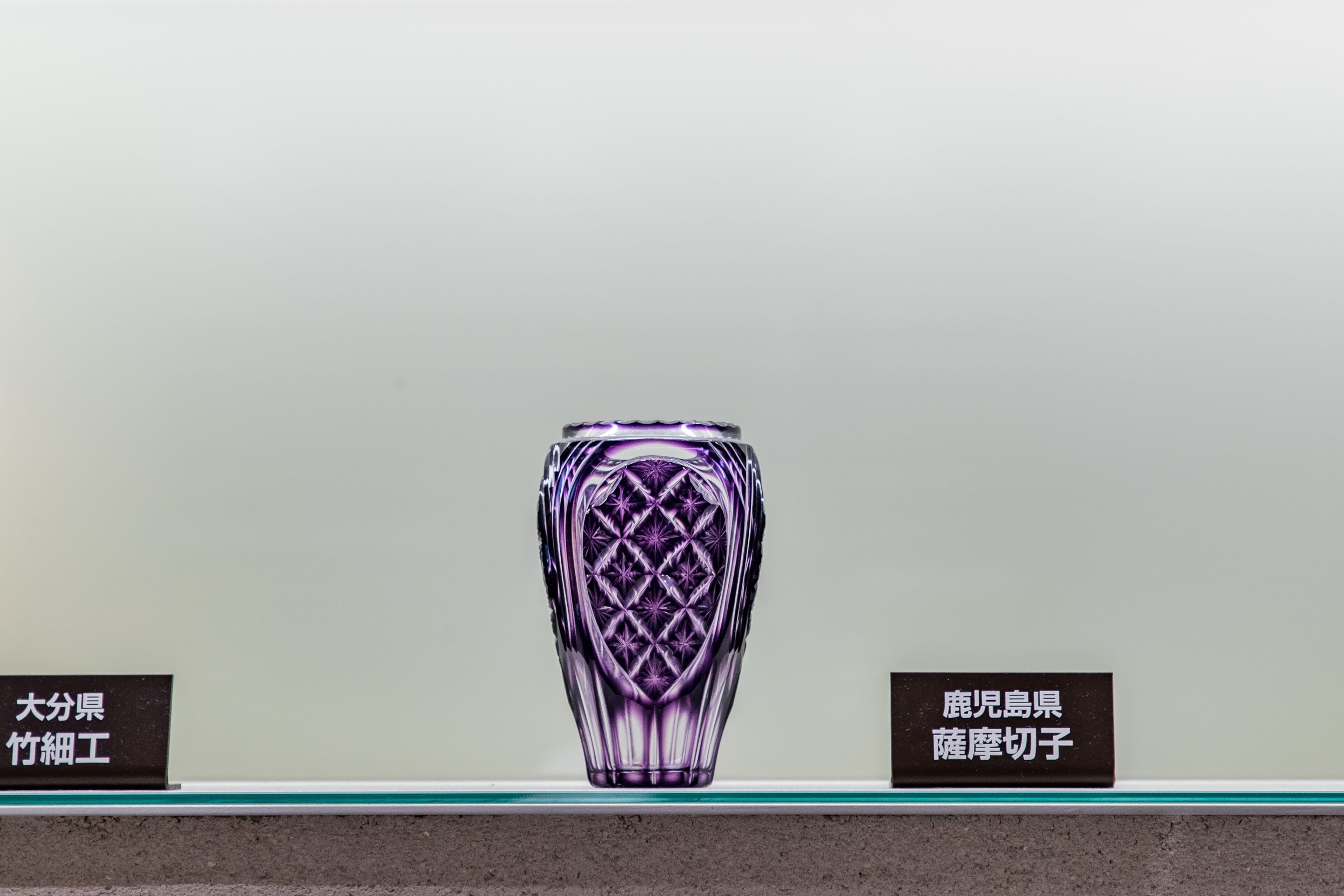 A purple glass vase with a geometric pattern, displayed on a shelf with two labels.