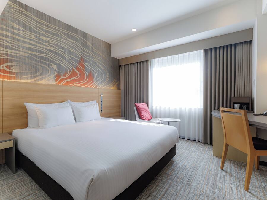 A hotel room with a large bed, a desk, a chair, and a view of the outdoors through the window.