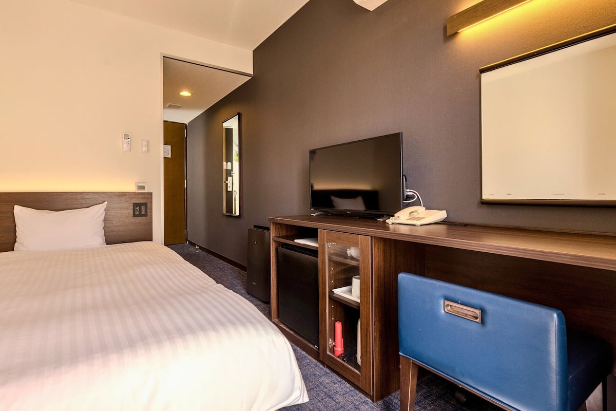 Una habitación de hotel con una cama, televisión, escritorio y teléfono, además de un espejo.